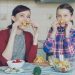 Joyful mother and cute cheerful daughter have fun grimacing silly with vegetables and looking into camera while cooking in the kitchen at home. Family, cook, and people concept